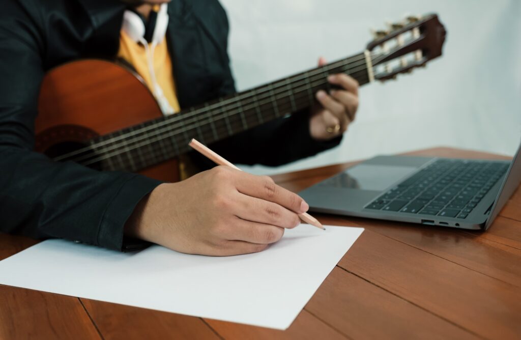 Young man musician hand holding a pencil to take notes of the music lesson, learning guitar online in the room with laptop.